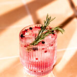 A gin and tonic cocktail with ice, rosemary, and peppercorns is served in a ribbed glass on a tiled surface.