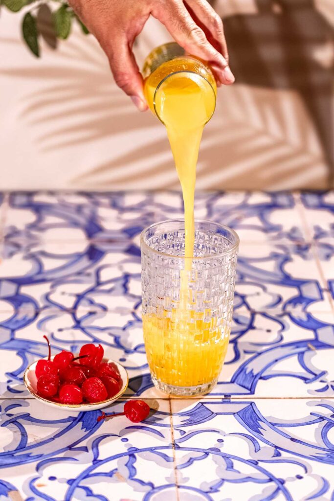 Orange juice poured into a cocktail shaker on a blue and white tiled surface.