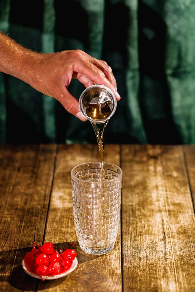 A person pours vermouth from a cup into an empty glass on a wooden surface near a small plate of cherries.