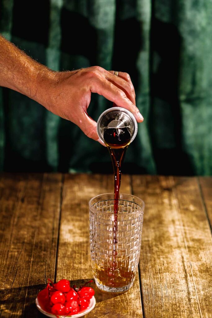 A hand pours whiskey from a small cup into a glass on a wooden table beside a bowl of cherries.