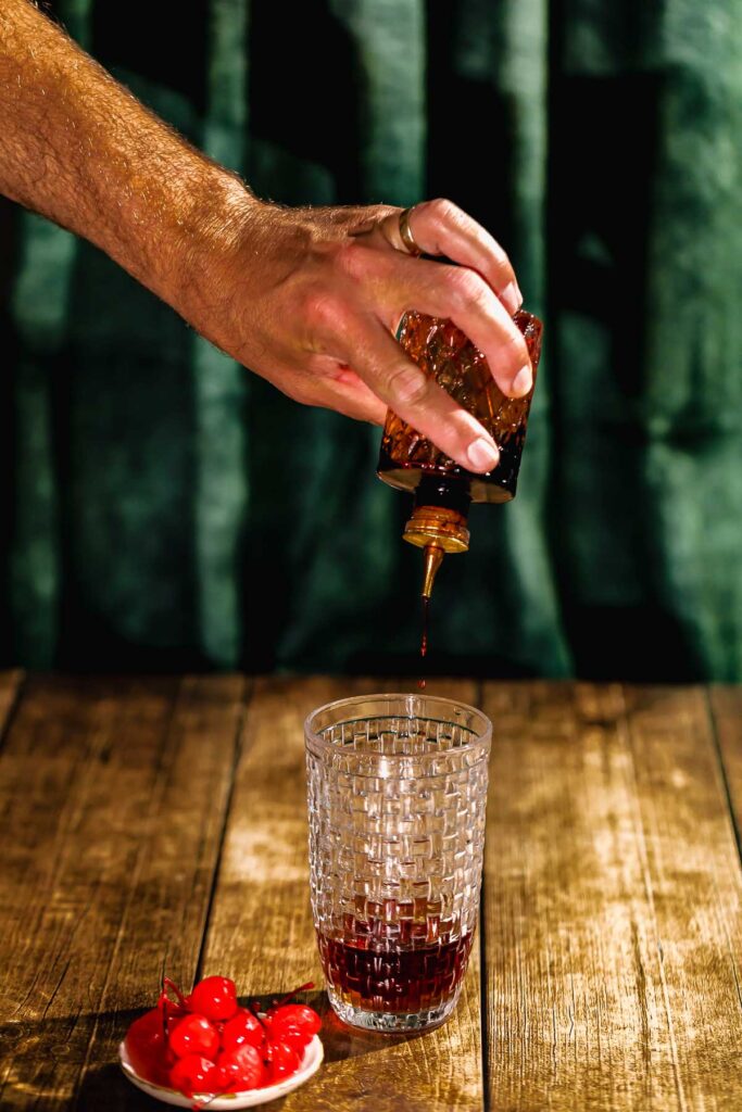 A hand pours bitters from a small glass bottle into a textured glass on a wooden table beside a bowl of cherries.