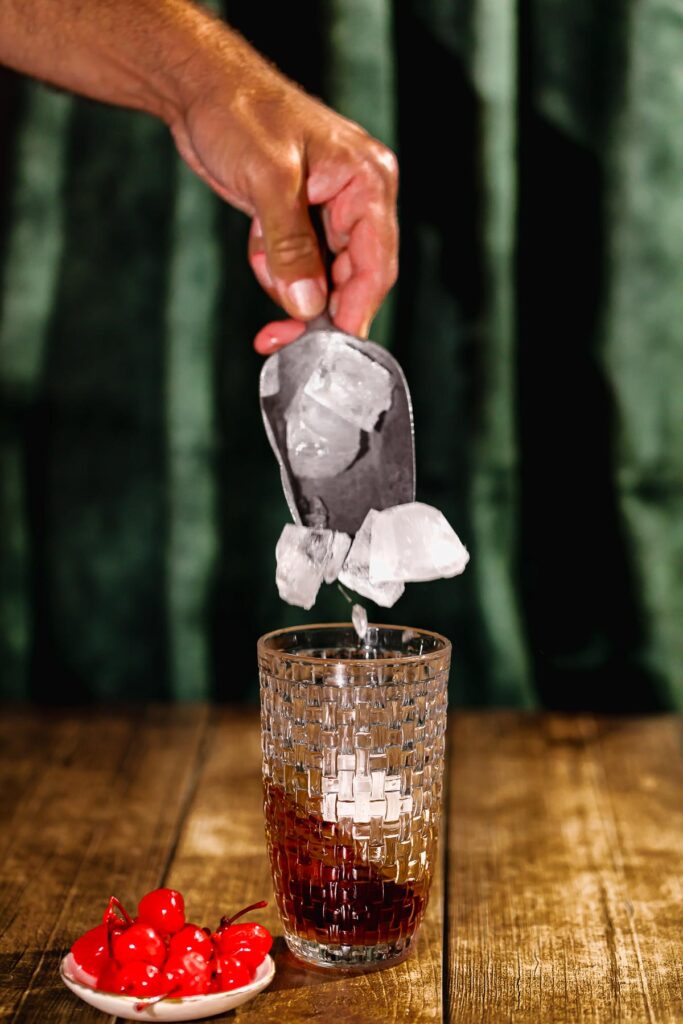 Ice cubes are being poured into a textured cocktail shaker of manhattan ingredients, with a plate of red cherries beside it on a wooden surface.