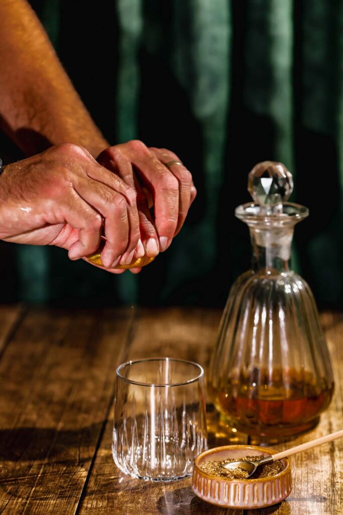 Hands squeeze an orange over a glass beside a decanter and container on a wooden table.
