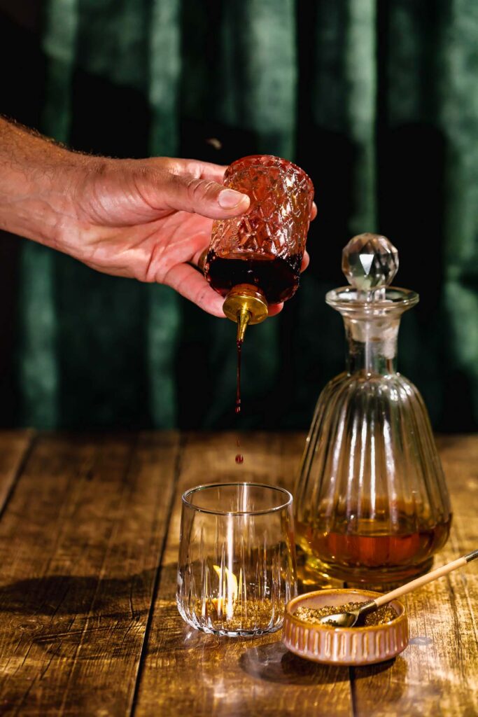 Dark bitters are poured from an ornate jar into a glass on a wooden table with a decanter of rum in the background.