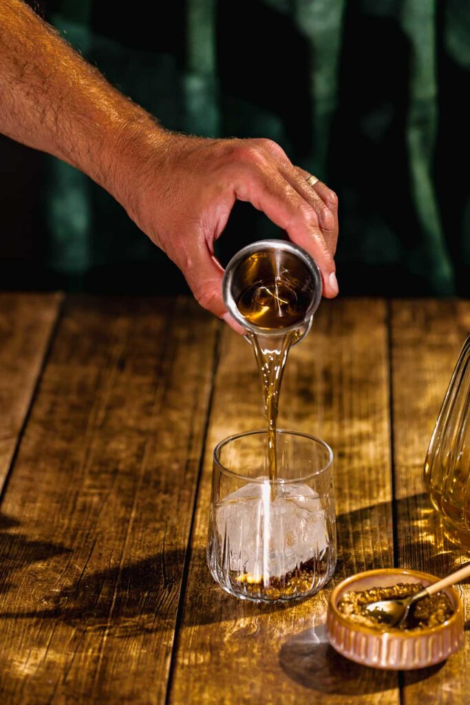 A hand pours rum from a jigger over ice in a glass on a wooden table.