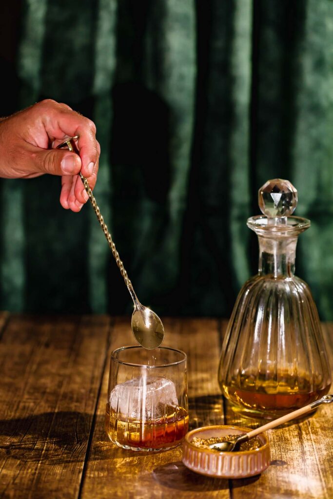 A hand stirs a rum old fashioned with ice in a glass using a spoon, while a decanter and lidded dish sit on a wooden surface.