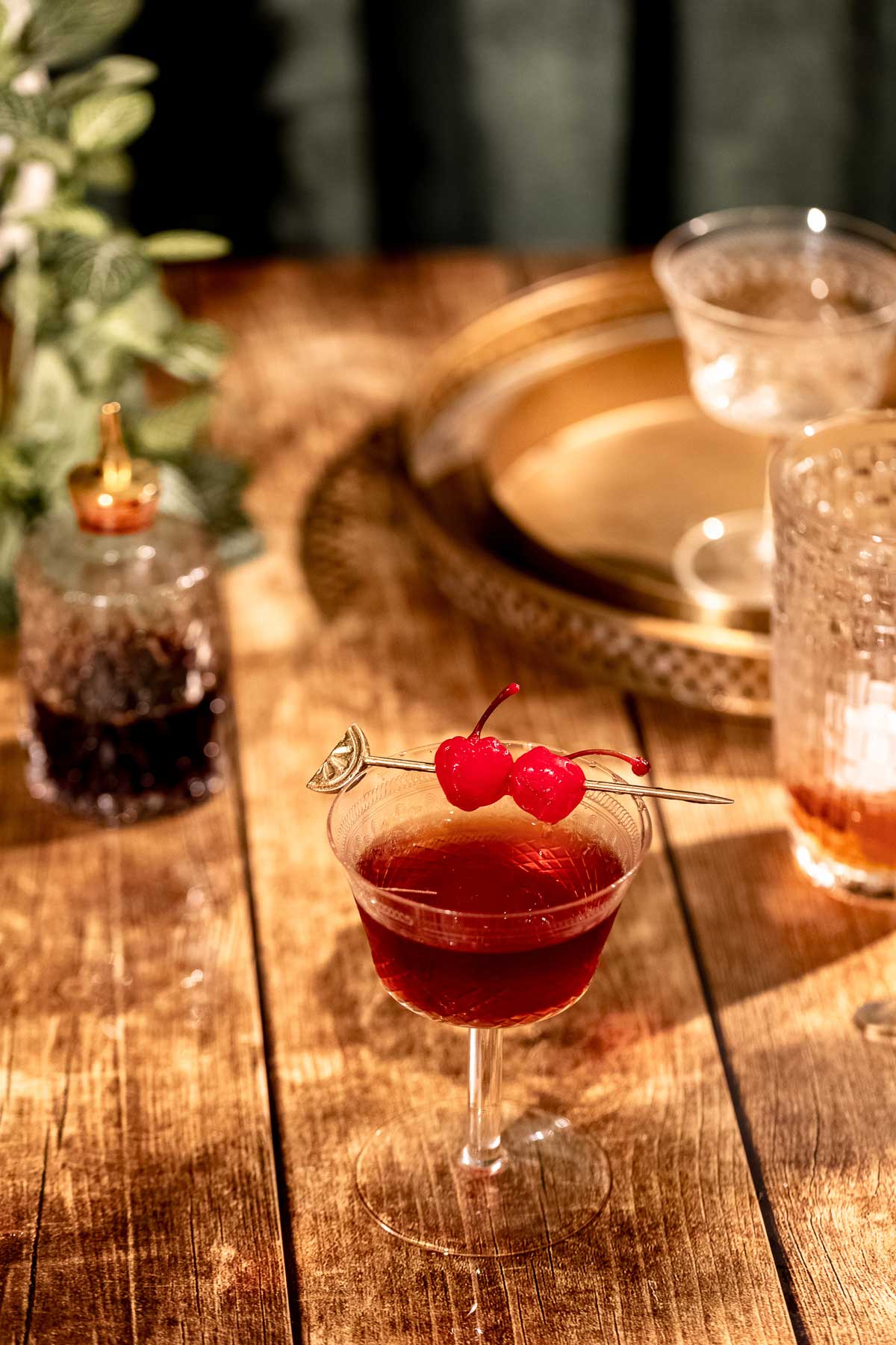 A Manhattan cocktail garnished with two cherries on a pick sits on a wooden table, with a gold tray and glassware in the background.