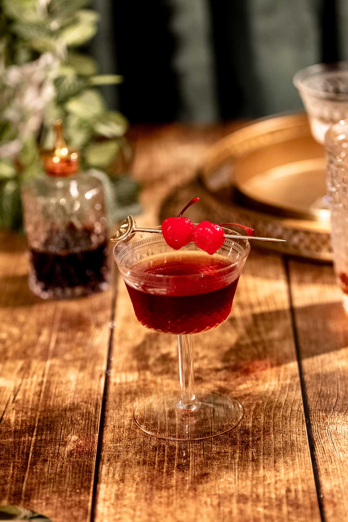 A Manhattan cocktail garnished with cherries sits on a wooden table with a blurred decanter and tray in the background.