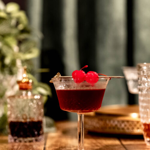 A stemmed cocktail glass filled with a Manhattan garnished with red cherries sits on a wooden table amidst drinks against a blurred green background.