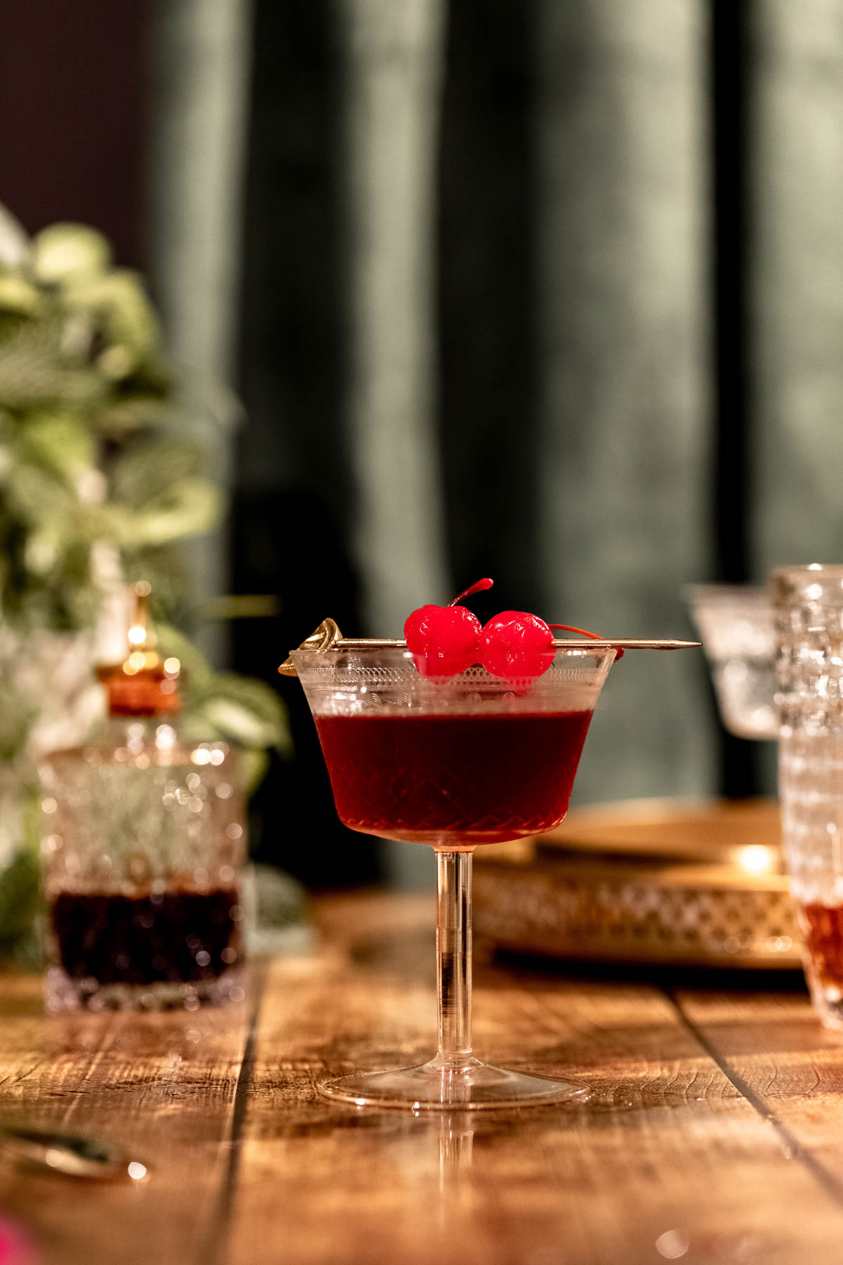 A stemmed cocktail glass filled with a Manhattan garnished with red cherries sits on a wooden table amidst drinks against a blurred green background.