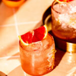 A paloma cocktail with ice and a red citrus slice garnish sits on a tiled surface.