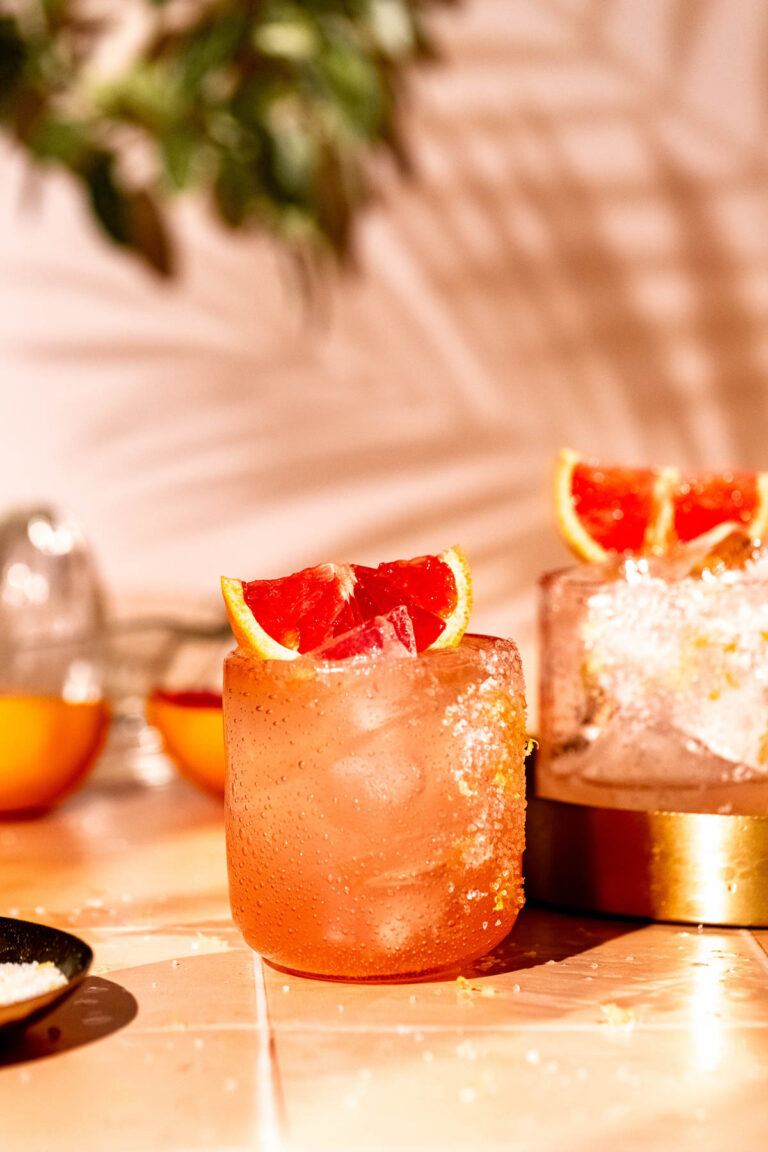 A pink paloma cocktail with ice and a slice of red citrus is on a table surrounded by scattered ice and citrus.