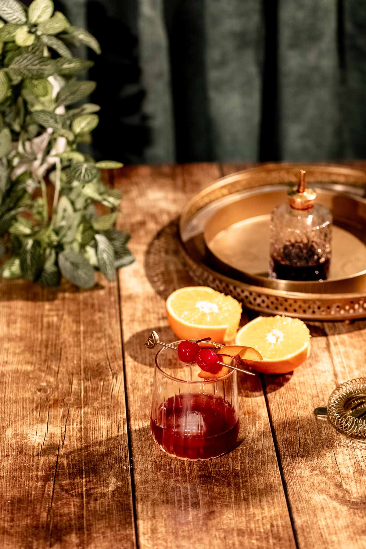 A glass with a rum old fashioned with two cherries on a skewer sits on a wooden table near halved oranges and a brass tray with a small bottle, set against a background of green plants.