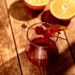 A glass of rum old fashioned cocktail with cherries and an orange peel on a skewer sits on a wooden surface, with two halved oranges in the background.