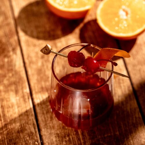 A glass of rum old fashioned cocktail with cherries and an orange peel on a skewer sits on a wooden surface, with two halved oranges in the background.
