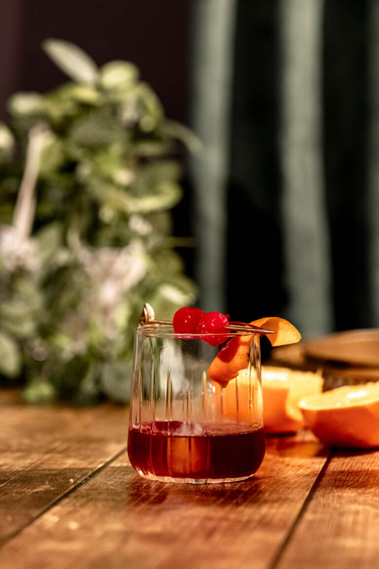 A rum old fashioned garnished with orange peel and cherries sits in a glass on a wooden table, set against blurred greenery.