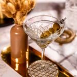 A clear vodka martini with olives rests on a gold tray beside a brown vase of dried flowers.