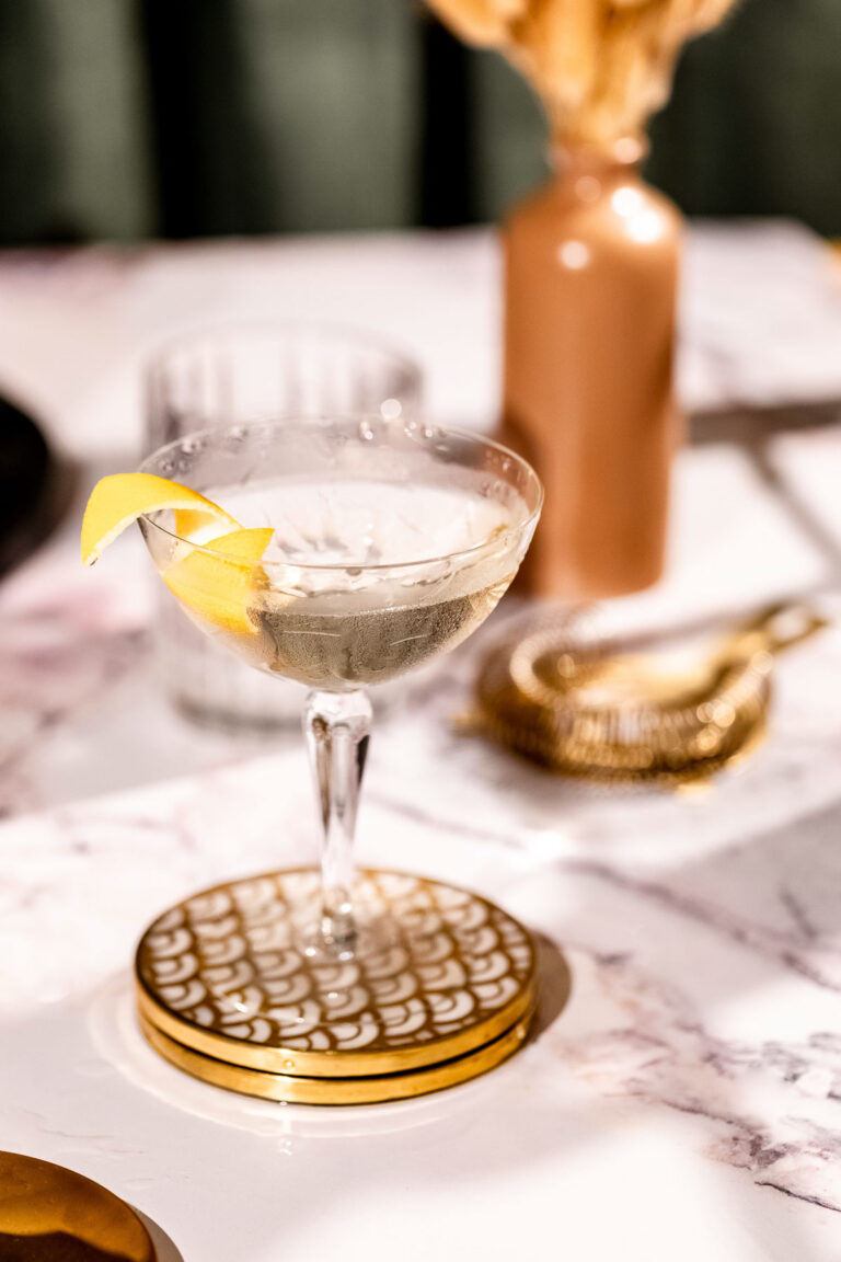 A lemon twist-garnished wet martini in a coupe glass sits on a gold-patterned coaster on a marble surface, with a glass, strainer, and vase in the background.