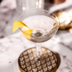 A wet martini cocktail garnished with a lemon twist sits on a decorative coaster on a marble surface, with a blurred glass in the background.