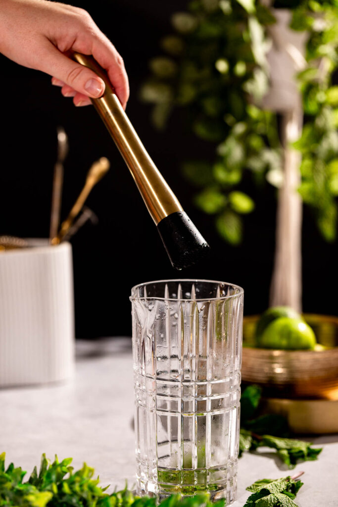 A hand muddles ingredients in a clear, patterned glass beside green leaves and a bowl of limes.