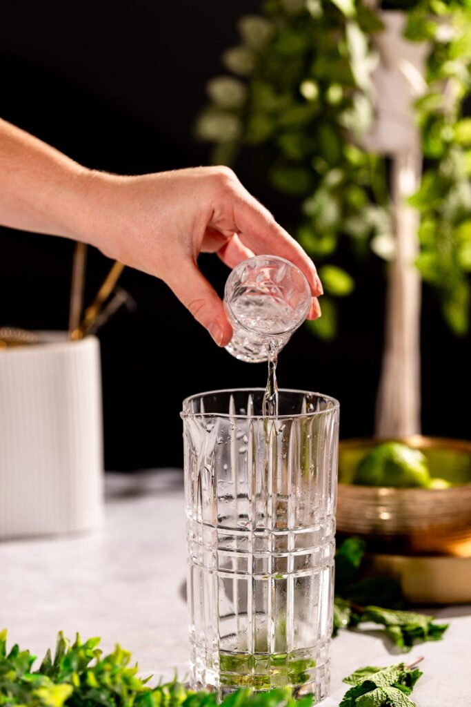 Rum is being poured from a small glass into a tall crystal glass, set against a backdrop of greenery and lime.