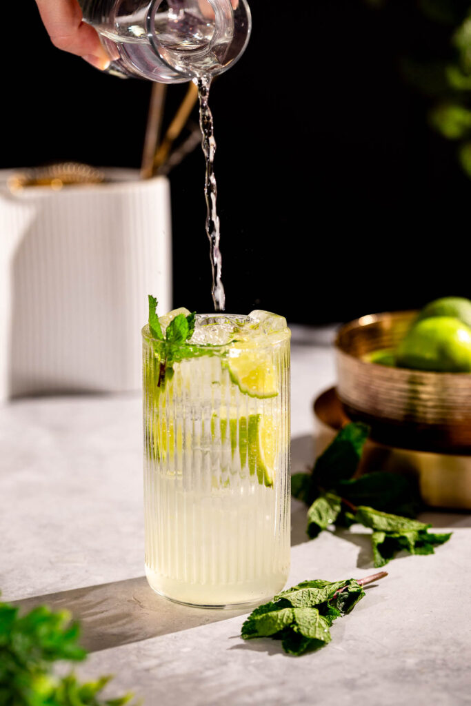 Soda water is being poured into a glass of iced lemonade with lime slices and mint, with mint leaves scattered on the table.
