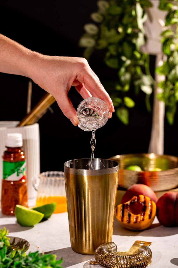 A hand pours tequila from a small glass into a metal shaker amid half a grilled peach, lime, strainer, syrup bottle, and greenery.