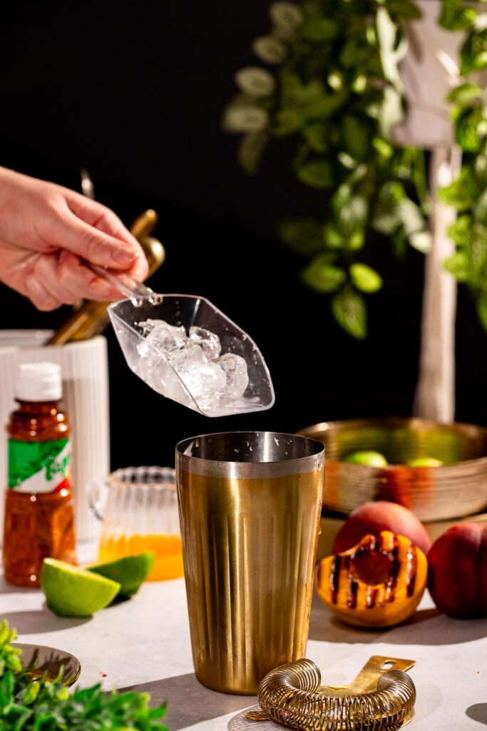 A person adds ice cubes to a gold cocktail shaker, with fresh peaches, a lime, and green leaves nearby.