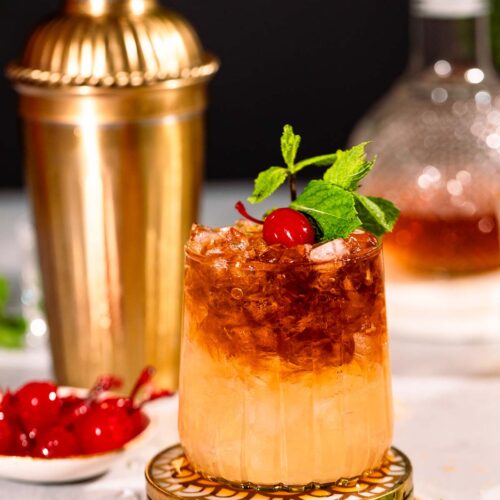 A mai tai cocktail with crushed ice, garnished with a cherry and mint sprig, sits in front of a gold shaker and scattered mint leaves and cherries.