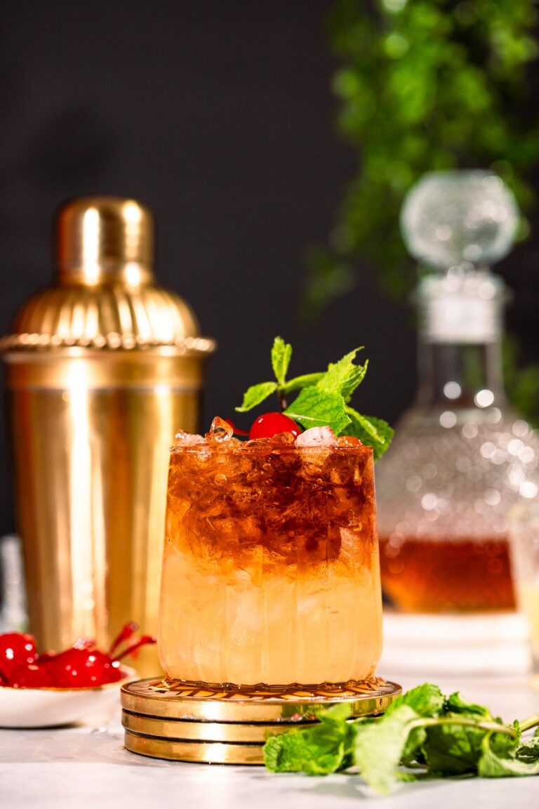 A mai tai cocktail garnished with mint and a cherry sits on a marble surface surrounded by cherries and mint, in front of a gold shaker and glass decanter.