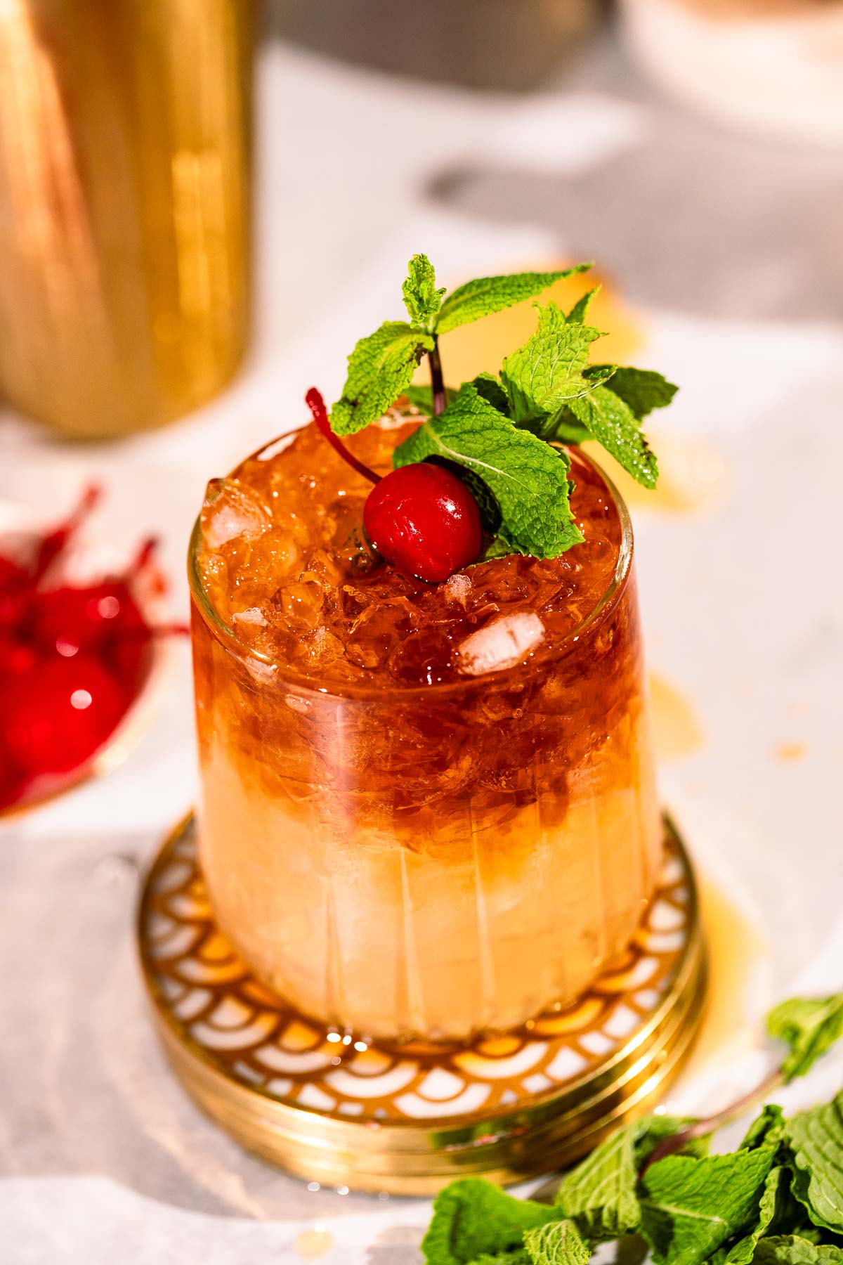 A layered mai tai cocktail topped with a cherry and mint sprig sits on a decorative coaster, surrounded by mint leaves and cocktail cherries.