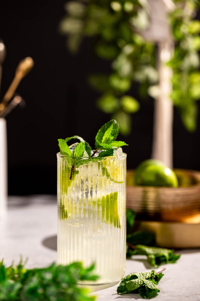 A mint mojito drink sits on a white surface, with green foliage and a bowl holding a lime in the blurred background.
