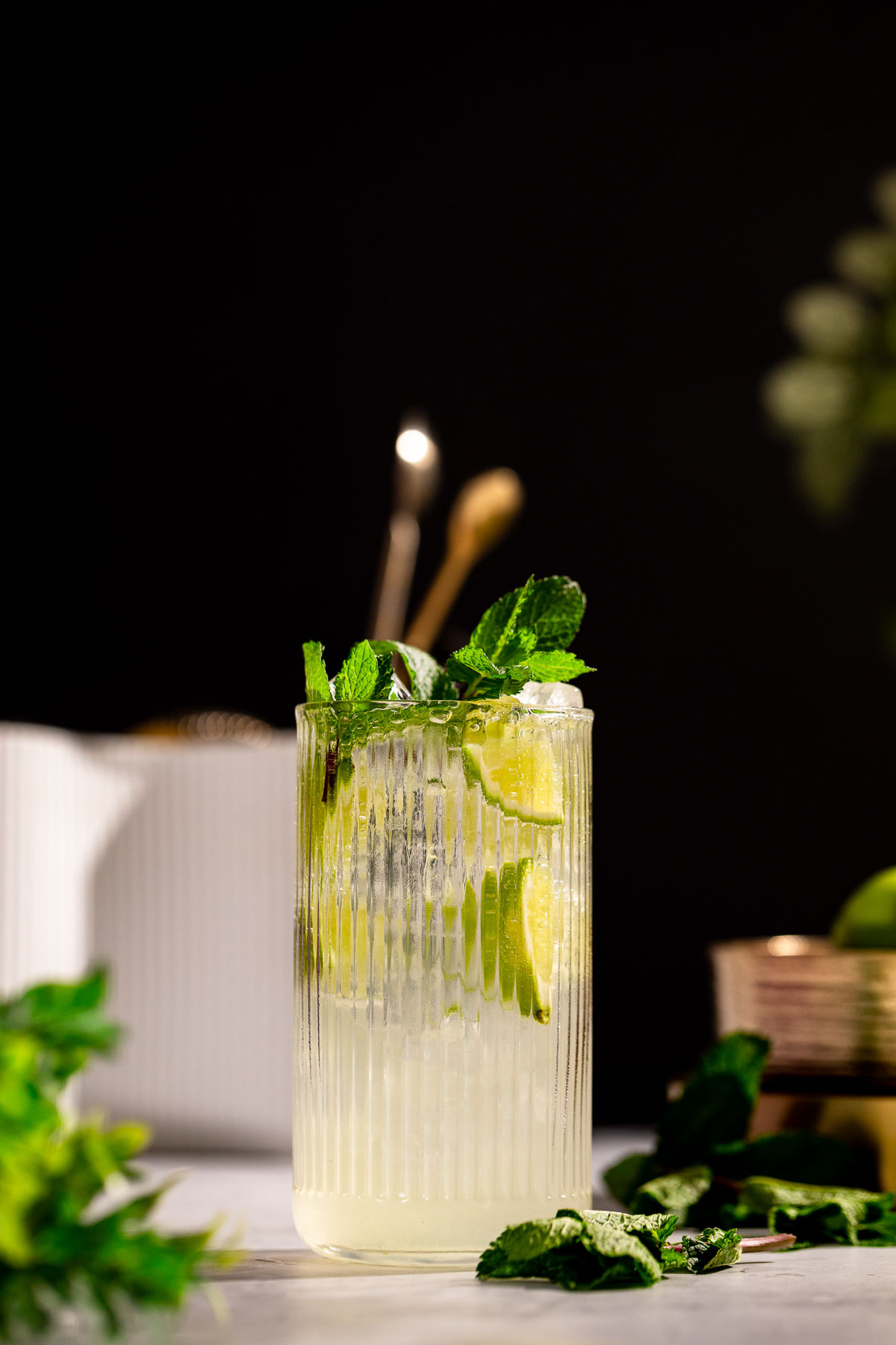 A tall textured glass holds a mint mojito cocktail with lime slices, mint leaves, and ice, surrounded by scattered mint sprigs on a dark background.