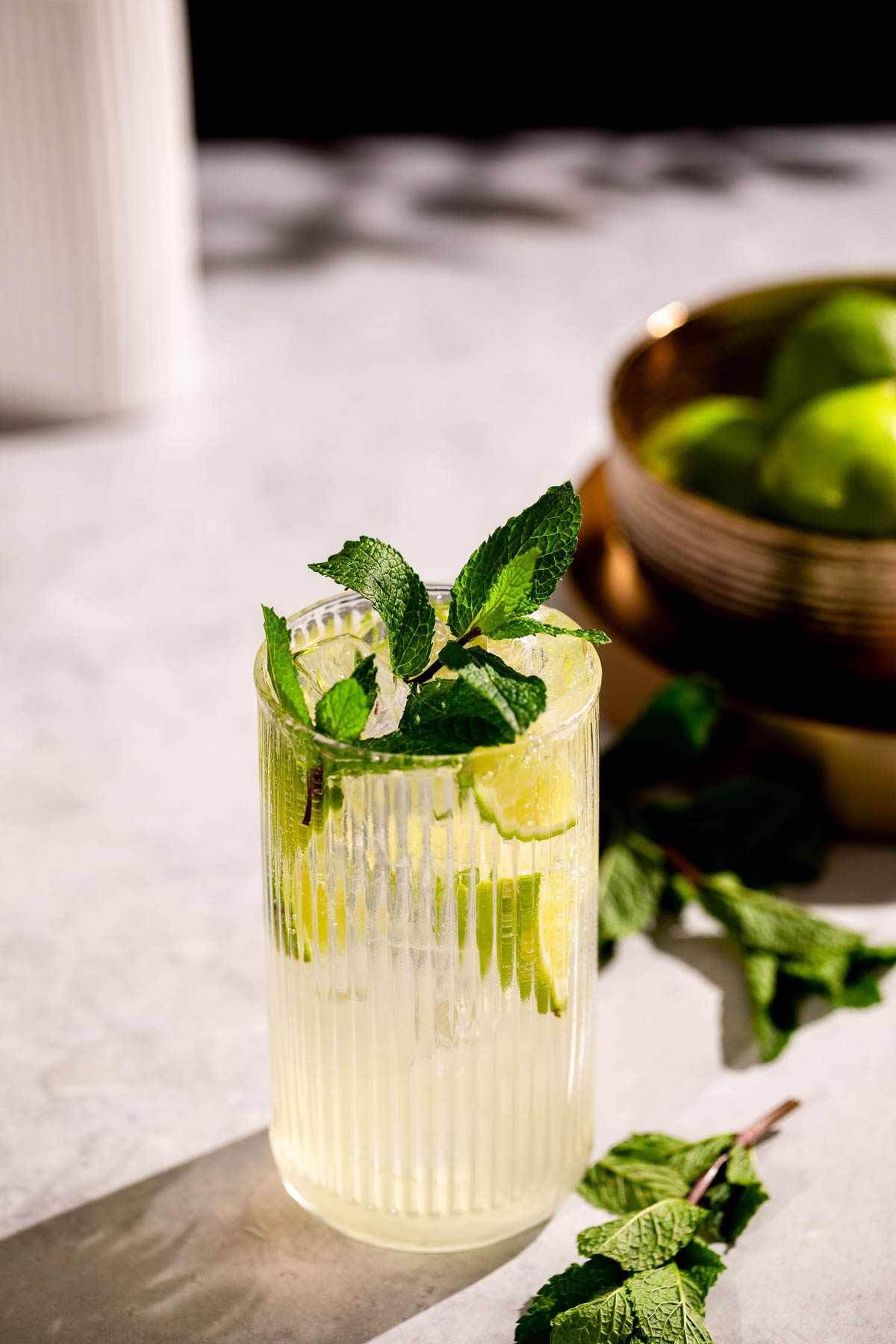 A tall glass of mojito with lime slices and mint leaves sits on a light surface with a bowl of limes in the background.