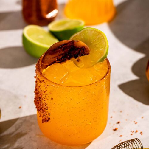 A grilled peach margarita with ice, garnished with a lime slice and chili powder on the rim, is set against a background featuring lime wedges and a bottle on a light surface.