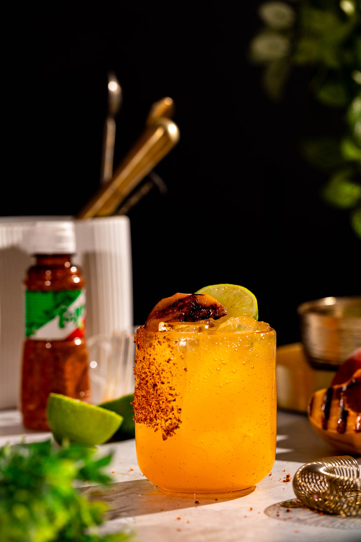 A grilled peach margarita with a chili powder rim, garnished with lime and charred fruit, sits near a spice bottle, lime slice, and kitchen utensils.