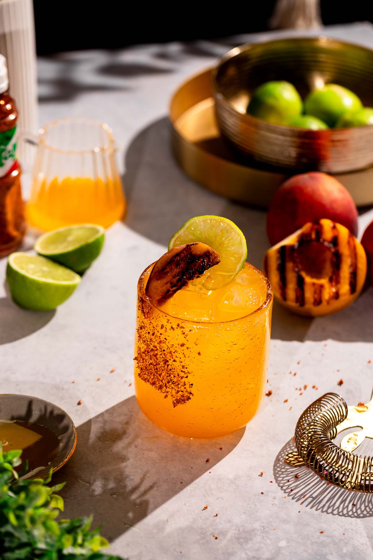 A glass of grilled peach margarita cocktail with a lime slice and grilled peach garnish sits on a gray surface, surrounded by limes, a peach, and a shaker.