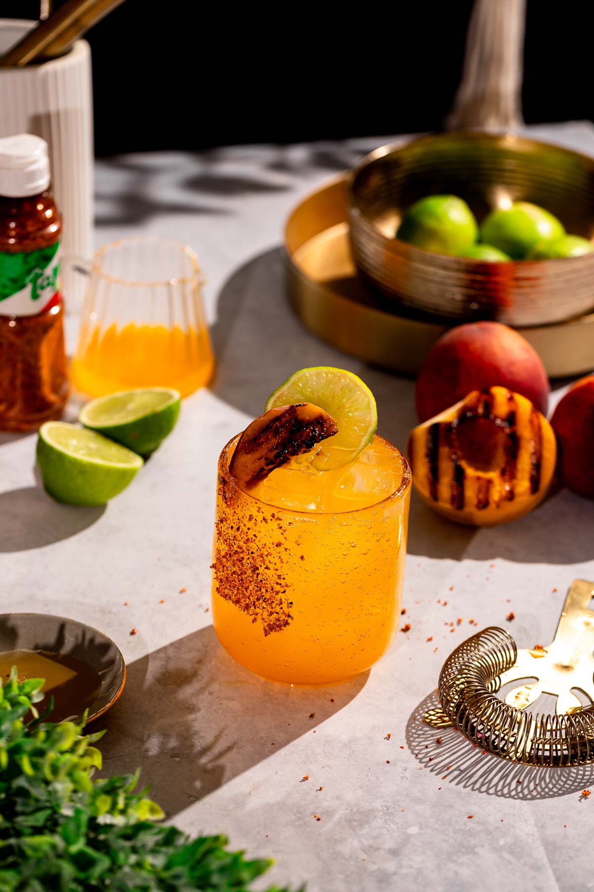 A cocktail glass with a grilled peach margarita drink garnished with lime and grilled peach sits on a table with limes, peaches, another drink, and a bottle of seasoning nearby.