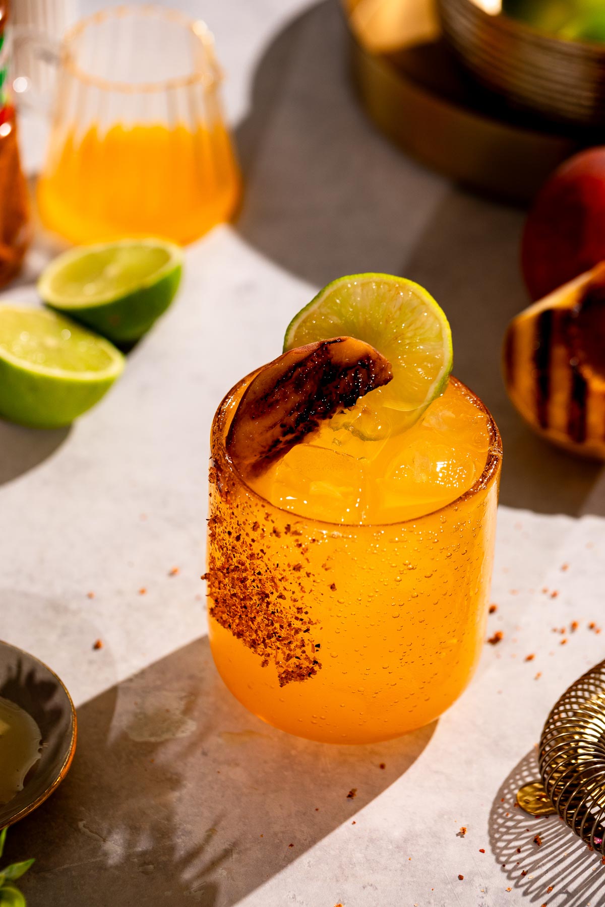 A grilled peach margarita cocktail garnished with a lime slice and grilled peach, on a chili-powdered rim, is set against a backdrop of lime halves and a glass of orange liquid.