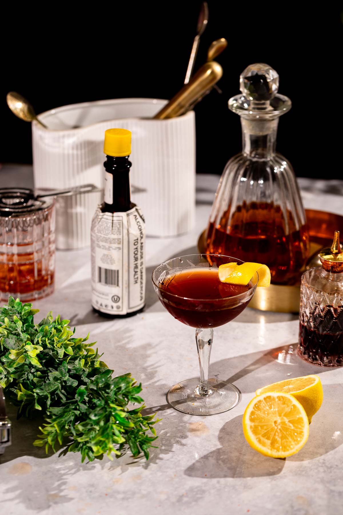 A Vieux Carré with a lemon twist is surrounded by a decanter, bitters bottle, sliced lemon, and bar tools on marble.