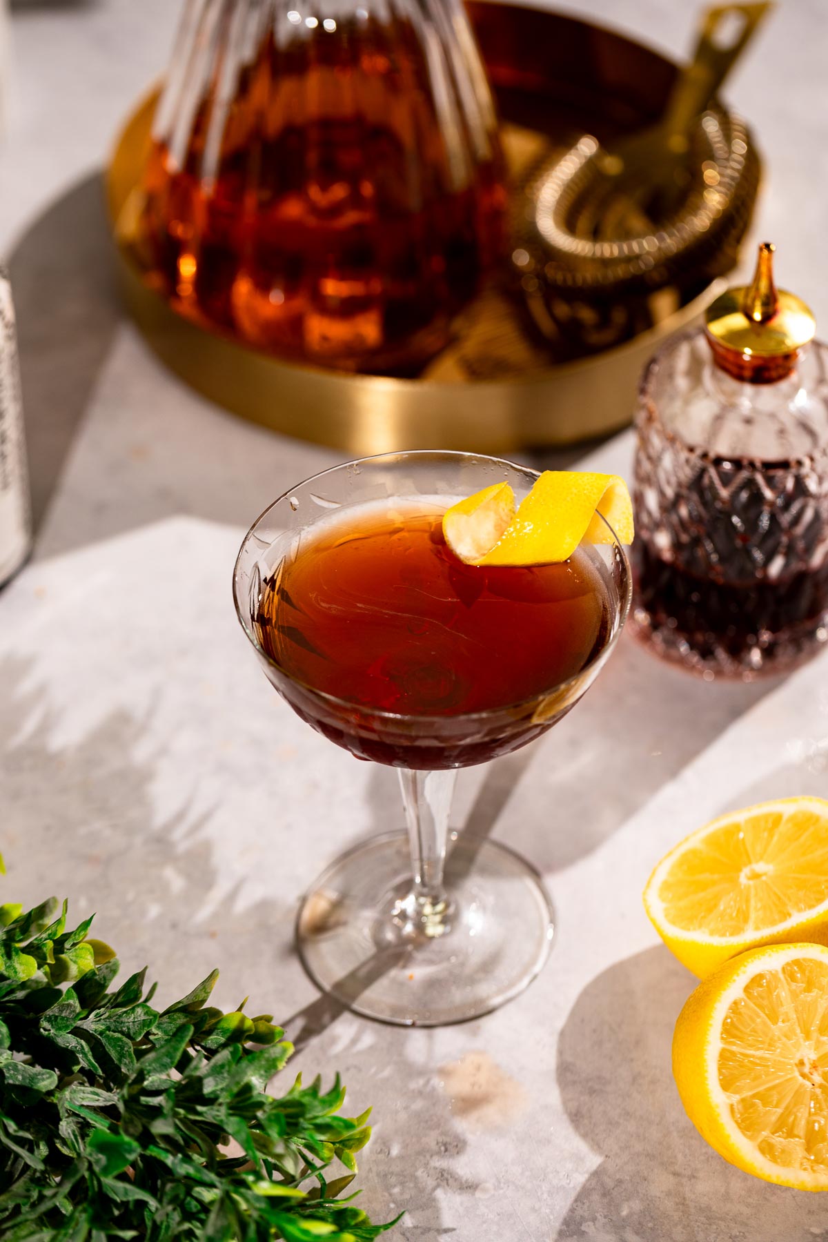 A Vieux Carré with a lemon twist sits on a surface next to a gold tray holding a decanter, bitters bottle, sliced lemons, and a small plant.