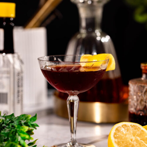 A coupe glass with a Vieux Carré cocktail and lemon twist garnish sits on a marble surface amid lush greens, a half lemon, and bottles.
