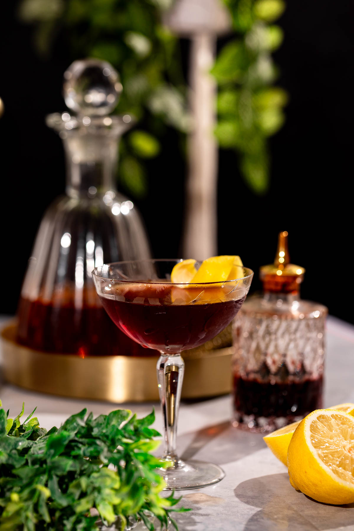 A Vieux Carré in a coupe glass, garnished with a lemon twist, sits on a table with a decanter, cut lemon, and greenery in the background.