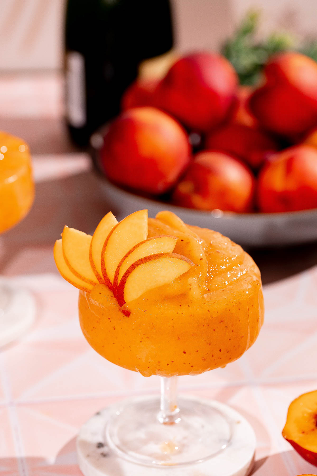 A slushy peach cocktail garnished with peach slices sits in a glass on a table with a bowl of peaches and a bottle in the background.