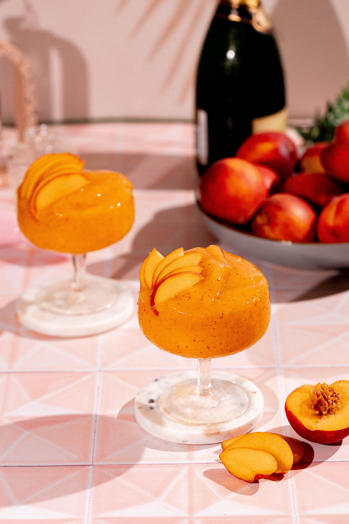Two peach bellini cocktails with peach slice garnishes sit on marble coasters, with a bowl of peaches and a champagne bottle in the background.