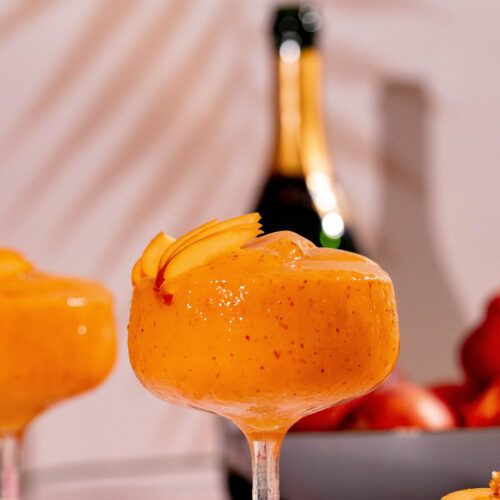 A frozen peach bellini cocktail garnished with slices sits in a glass, accompanied by a champagne bottle and a nearby bowl of peaches.