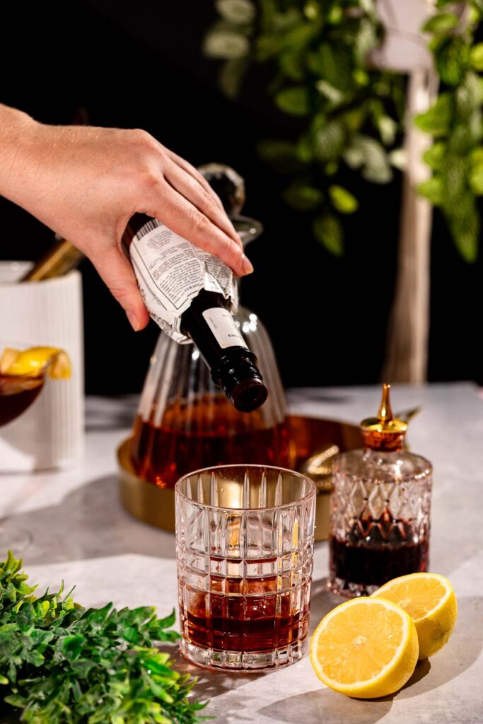 A hand pours Peychaud's bitters from a bottle into a glass on a table adorned with a decanter, lemon slices, a small stoppered bottle, and greenery.