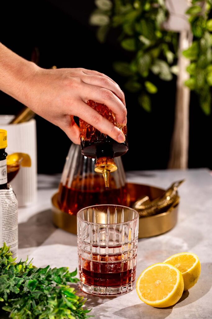 A hand pours bitters from a bottle into a glass with sliced lemons, greenery, and a container nearby on the countertop.