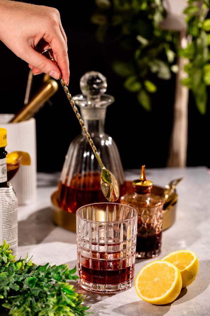 A hand stirs a drink in a glass with a metal spoon, surrounded by sliced lemons, a decanter, bitters bottle, and small jar, against a backdrop of greenery.