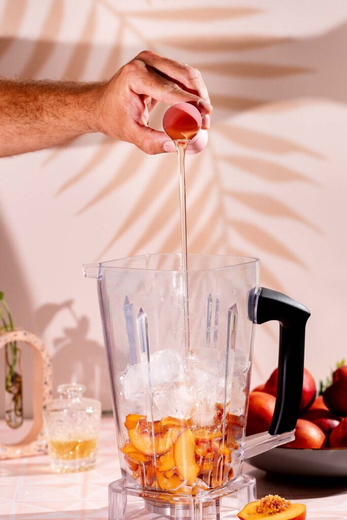 Simple syrup is poured from a small cup into a blender with ice and peach slices, while additional peaches sit on the countertop.